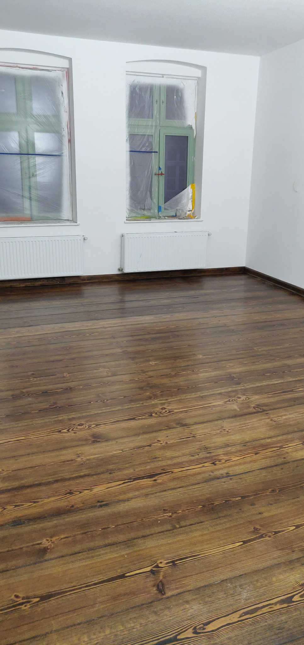 Odnowiona, lakierowana podłoga z desek w jasnym pomieszczeniu z dwoma oknami zabezpieczonymi folią malarską.