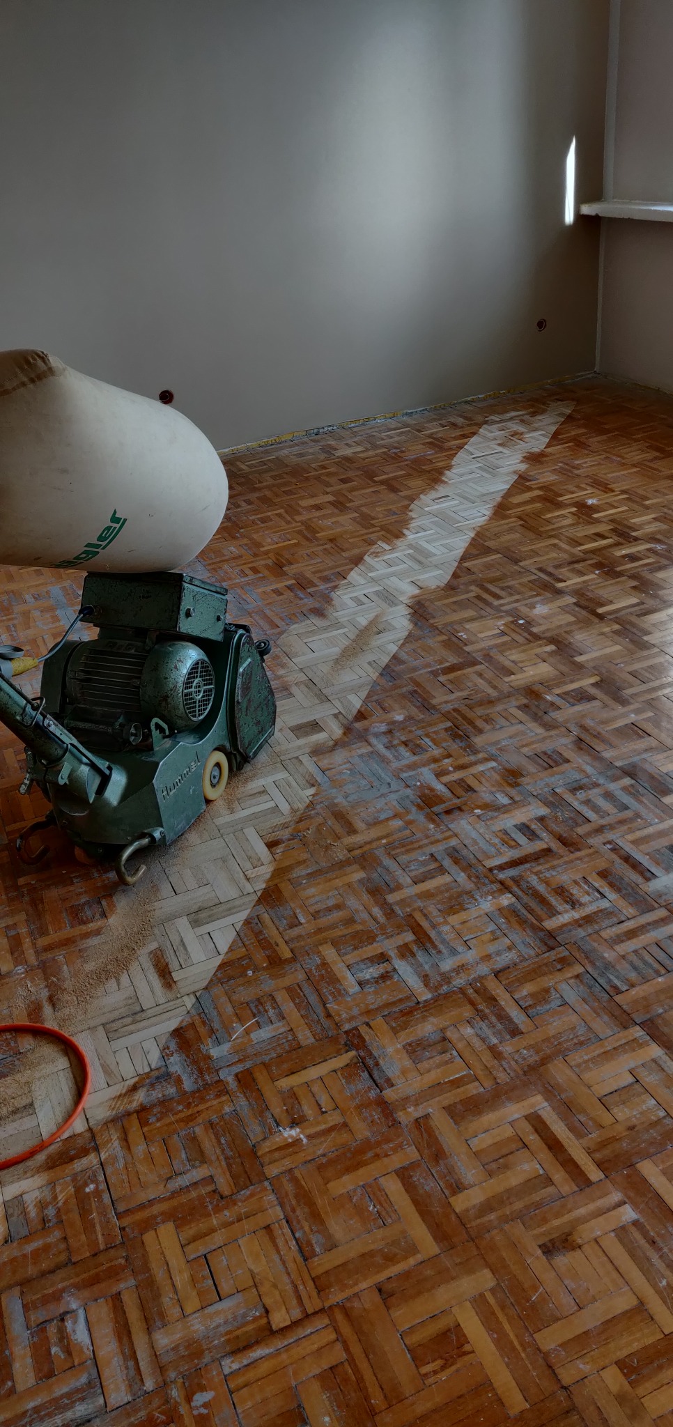 Parkiet w jodełkę częściowo wyszlifowany, widoczna maszyna do szlifowania parkietu marki Hummel i worek na pył.