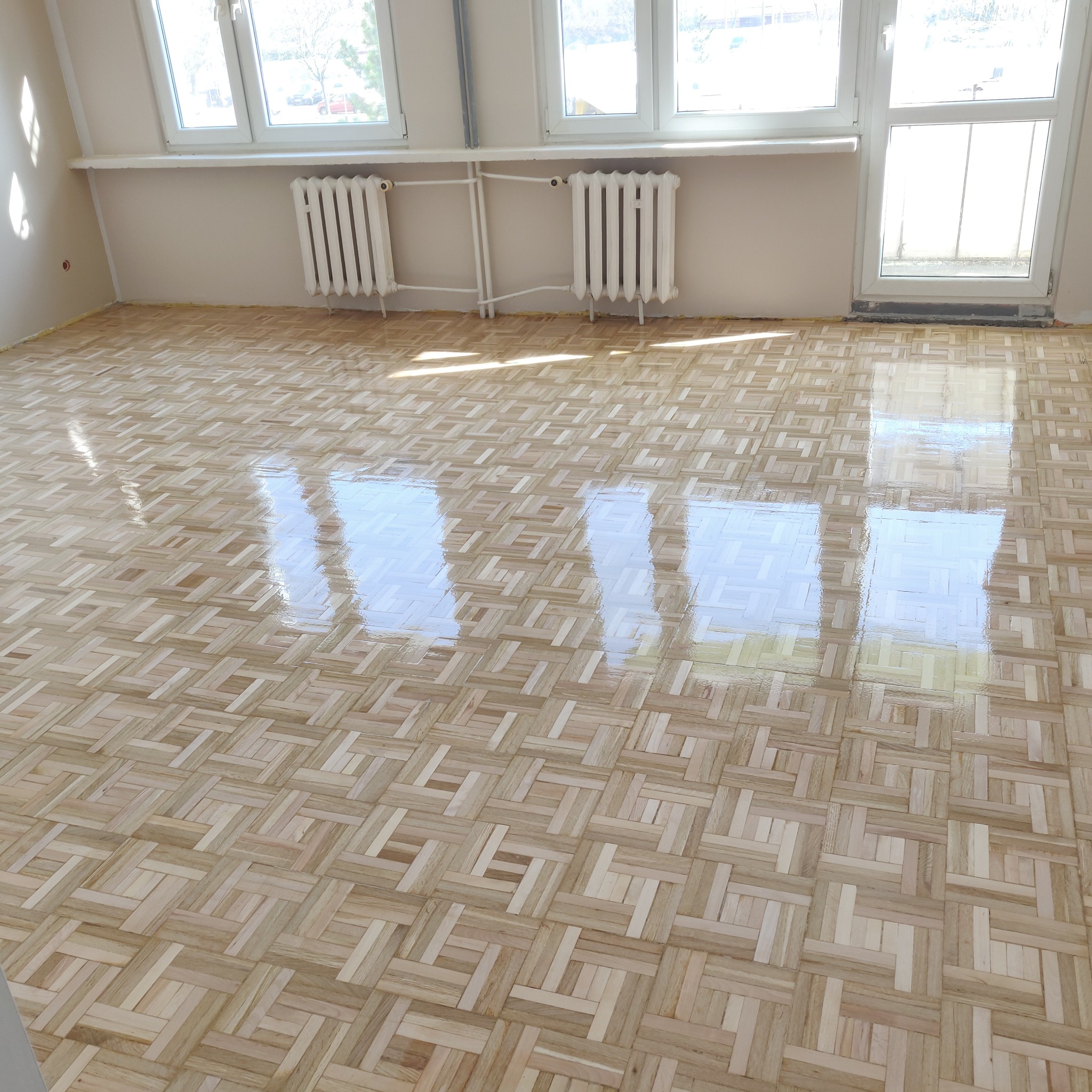 Błyszcząca, drewniana podłoga ułożona w parkiet z wzorem koszykowym po renowacji, z widocznymi refleksami światła słonecznego wpadającego przez okna.