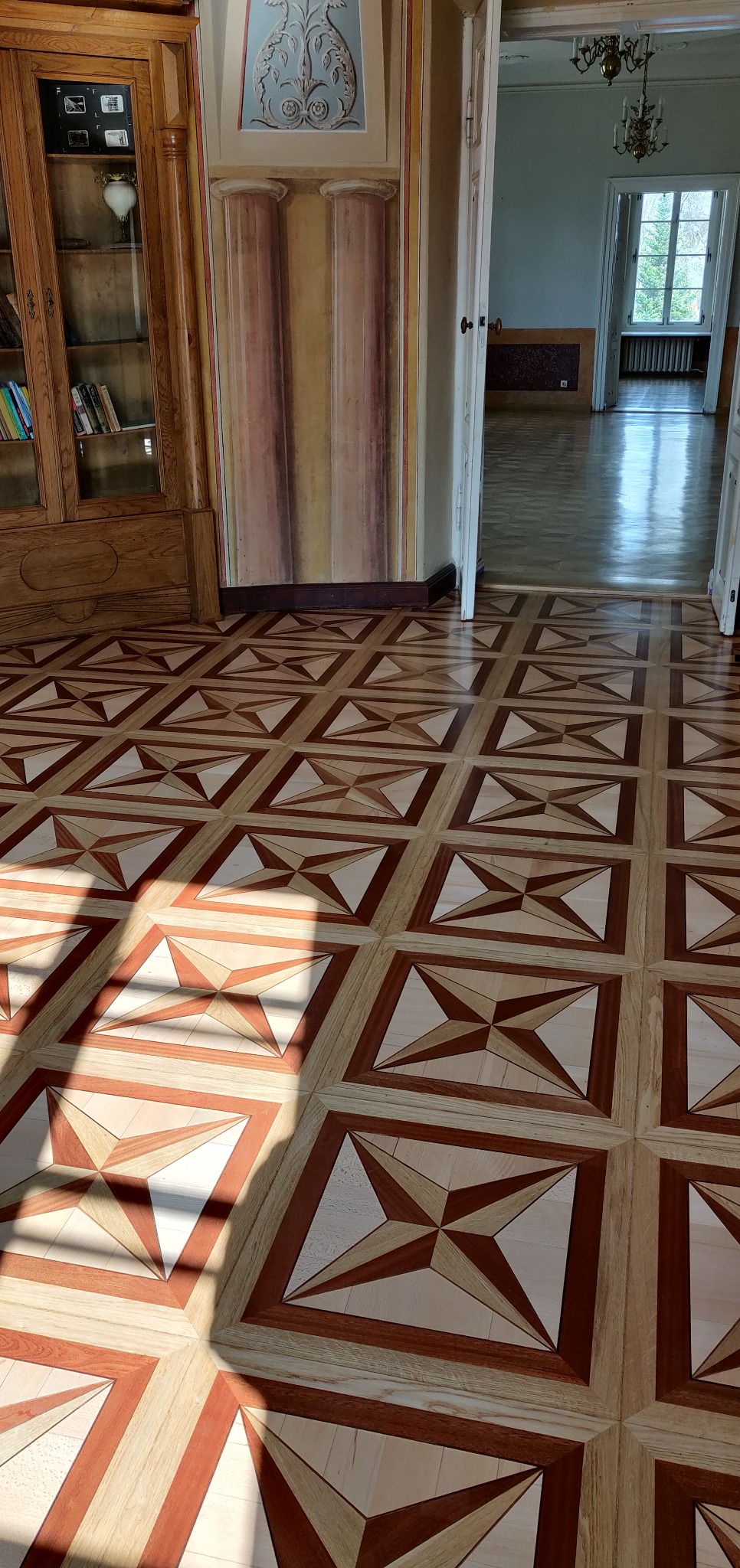 Wzór parkietu z jasnego i ciemnego drewna ułożony w geometryczne kwadraty i trójkąty, wpadające promienie słoneczne tworzą cień na podłodze, w tle widoczny regał z książkami i otwarte drzwi...