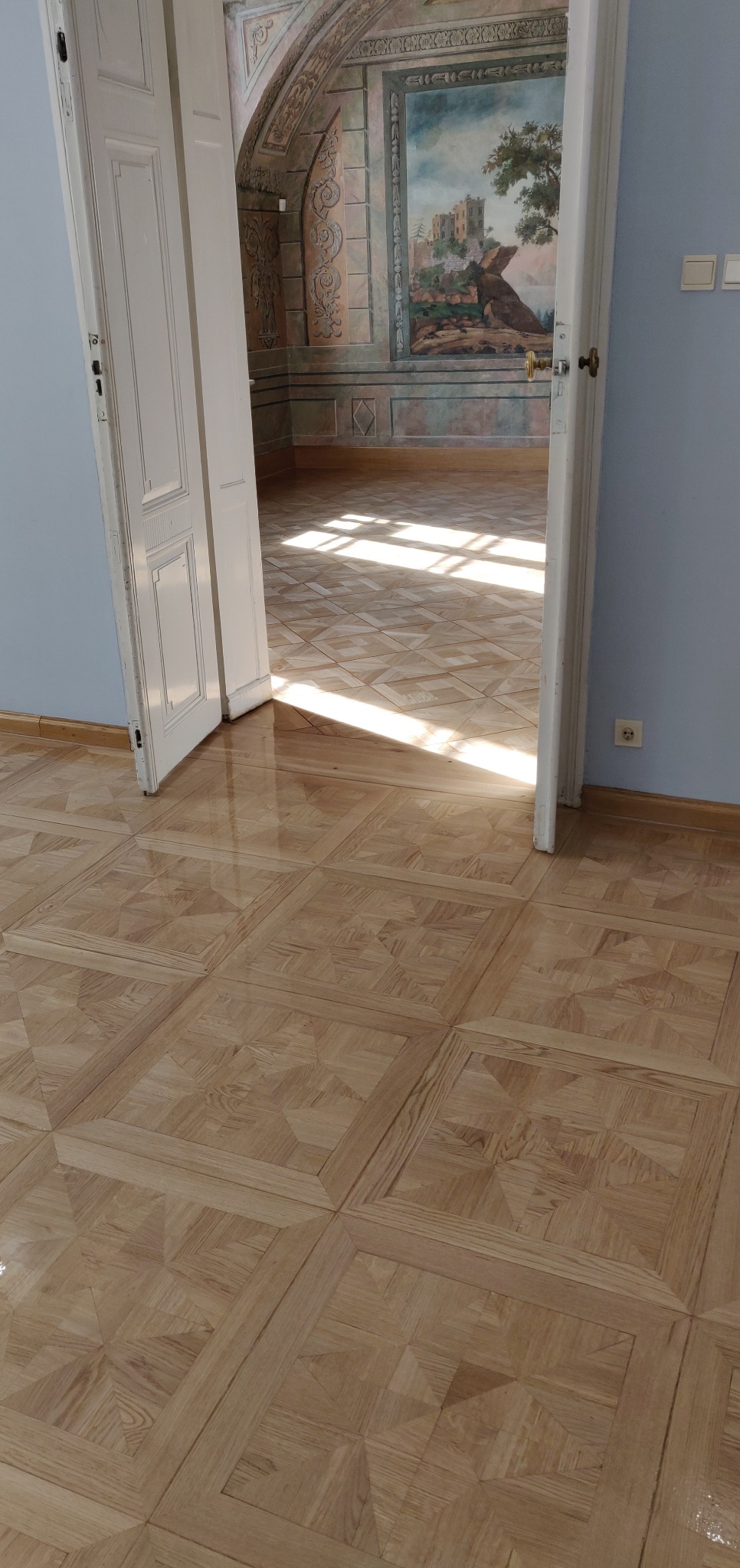 Parkiet dębowy ułożony we wzór kwadratów, świeżo polakierowany, widoczny w otwartych drzwiach z dekoracyjnym malowaniem na ścianie w tle, z refleksami słonecznego światła.