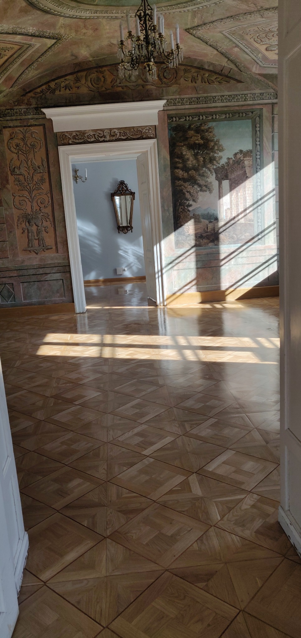 Parkiet ułożony we wzór rombów w przestronnym, odnowionym wnętrzu pałacowym, z promieniami słońca wpadającymi przez okno, oświetlającymi podłogę i malowidła na ścianach.
