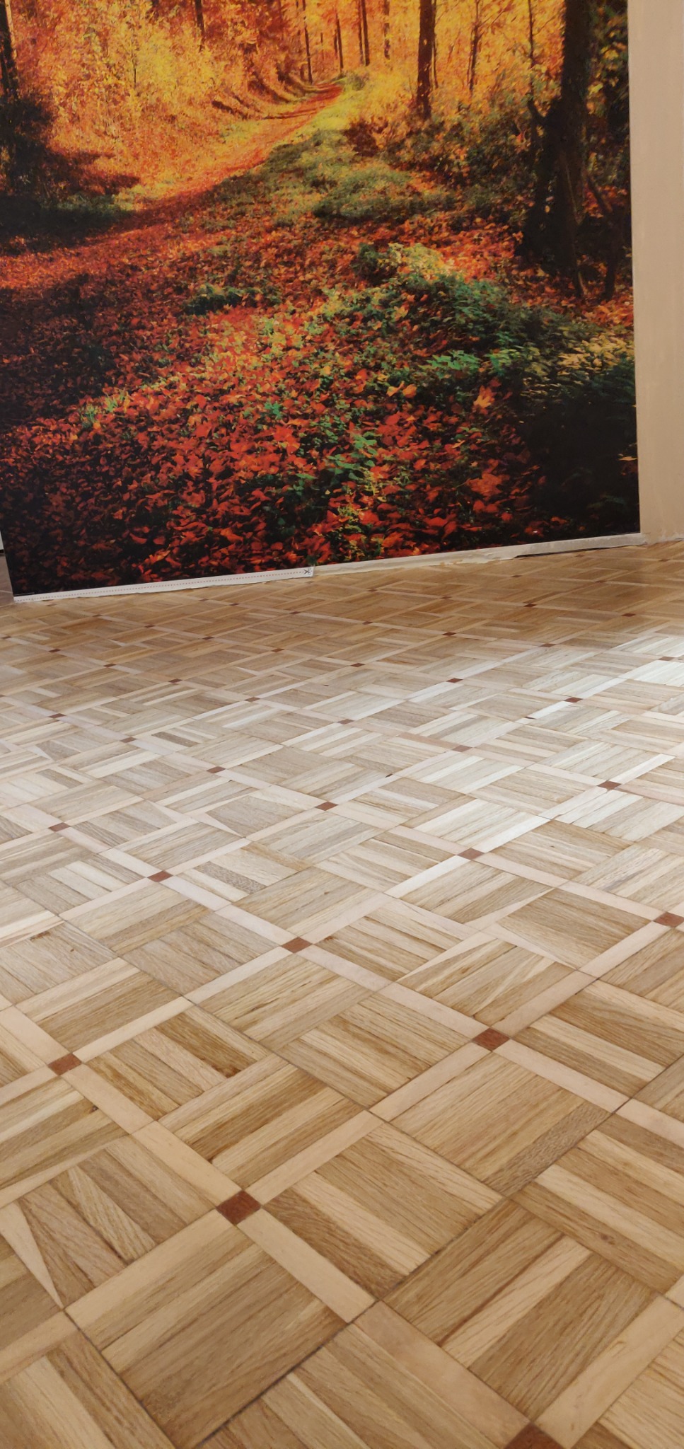 Parkiet ułożony we wzór szachownicy z jasnego drewna, z ciemniejszymi akcentami. W tle fototapeta z jesiennym lasem, tworząca ciepłą atmosferę wnętrza.
