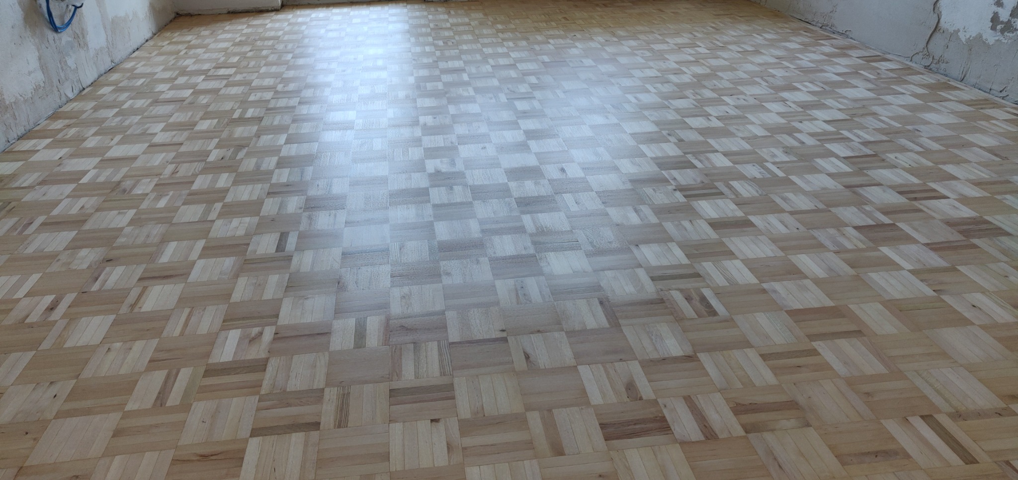 Parkiet ułożony w kwadraty, po cyklinowaniu, z widocznym odbiciem światła. Ściany w stanie surowym, przygotowane do dalszych prac wykończeniowych.
