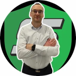 Mężczyzna w okularach, z zegarkiem na ręku, stoi na tle logotypu w odcieniach zieleni i szarości, prezentując się profesjonalnie.