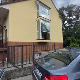 Elewacje-mycie-malowanie. - Jasnożółta elewacja domu z dwoma wykuszami, z których dolny ma pękniętą szybę w kształcie gwiazdy. Przed domem ciemnoszary samochód z hakiem holowniczym i częściowo widoczna przyczepa.
