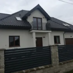 Nowoczesny dom jednorodzinny z ciemną dachówką i balkonem, ogrodzony grafitowym płotem z elementami imitującymi kamień.