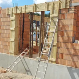 Wznoszone ściany z pustaków ceramicznych, drewniane szalunki, dwie aluminiowe drabiny na placu budowy w słoneczny dzień.