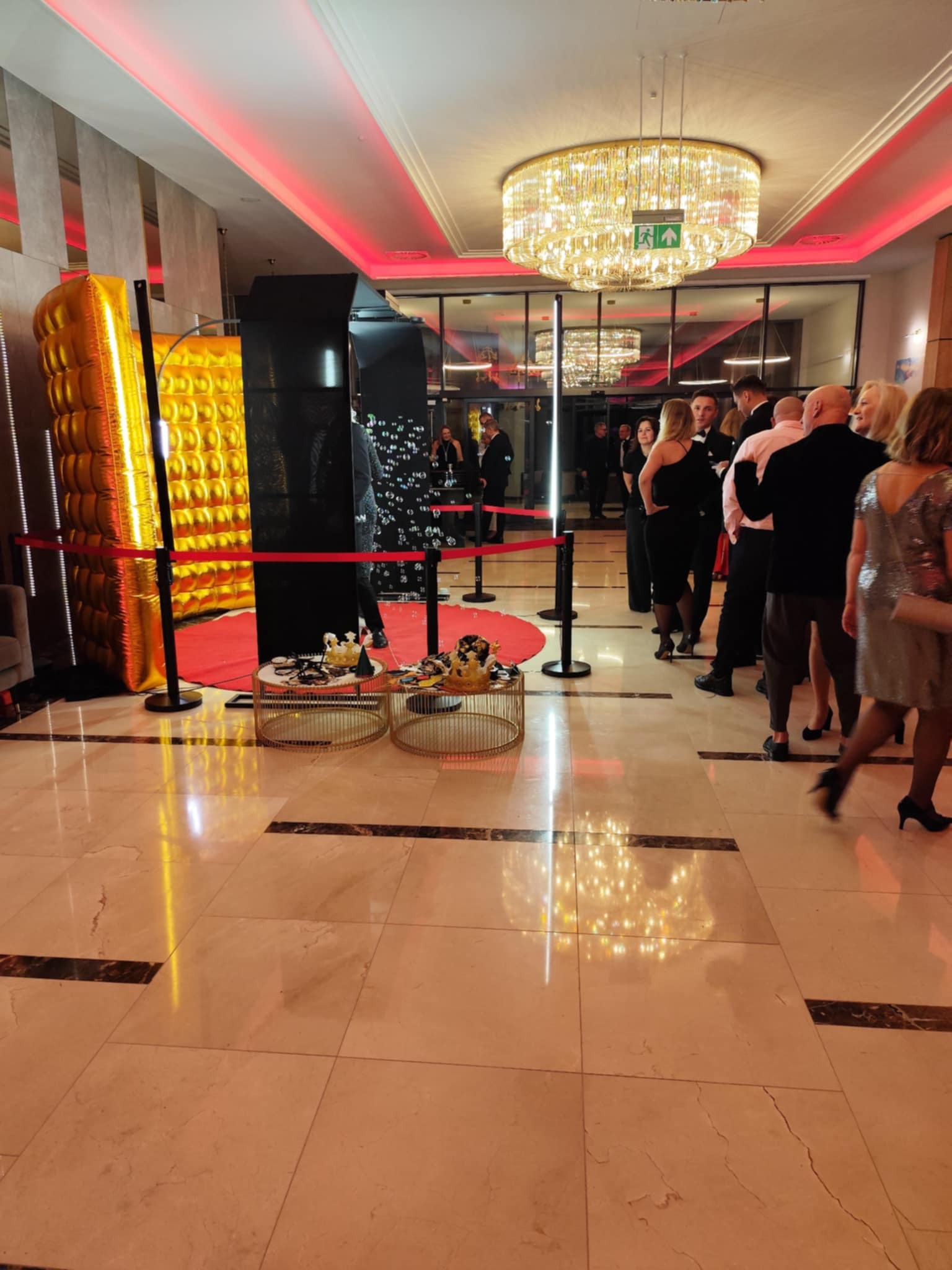 Czarno-złota fotobudka z rekwizytami na stolikach i czerwonym dywanem, ustawiona w eleganckim holu z oczekującymi gośćmi i kryształowym żyrandolem.