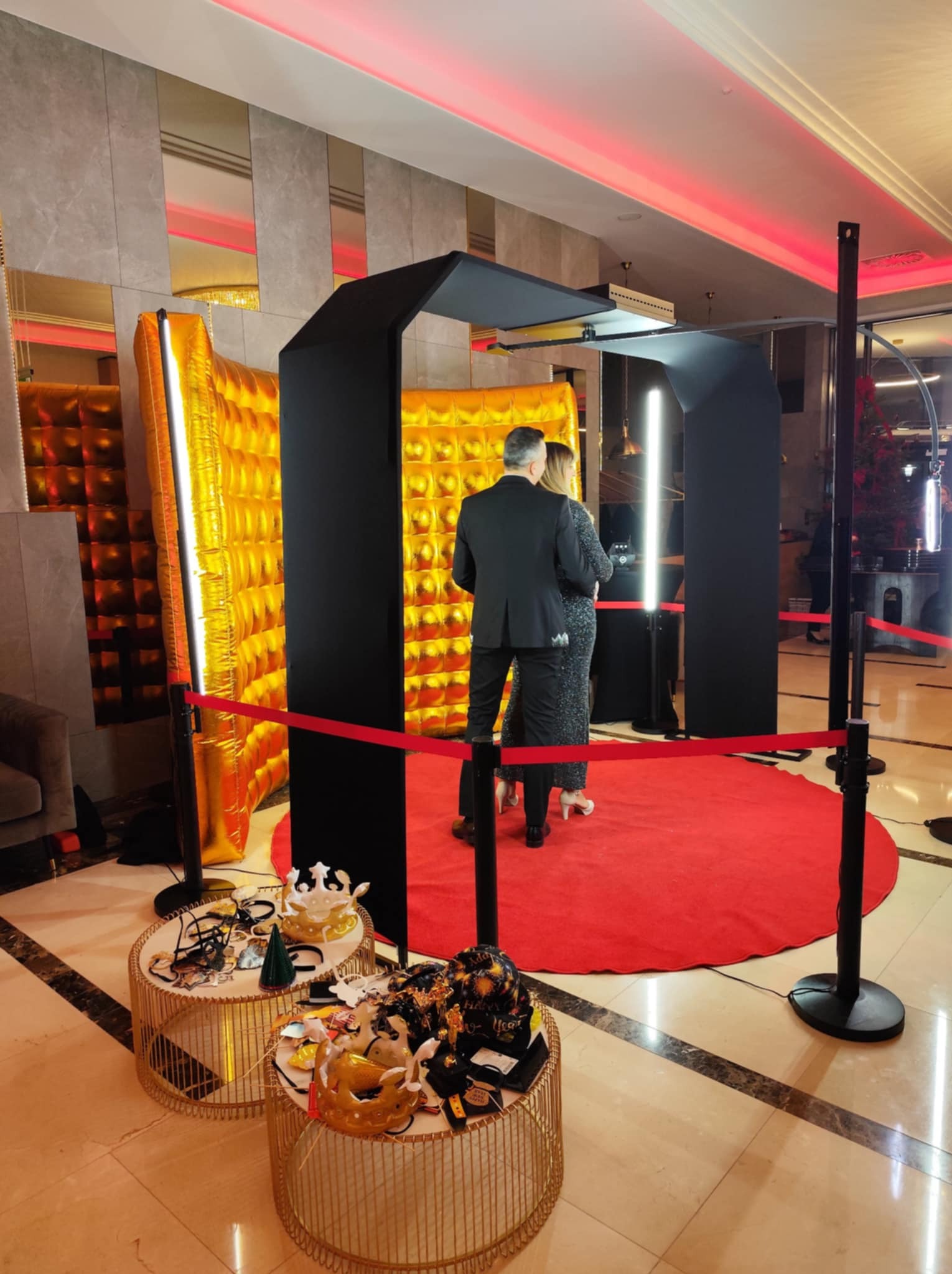 Para pozuje w fotobudce z czarną ramą i złotym tłem, na czerwonym dywanie. Stoliki z rekwizytami do zdjęć. Oświetlenie sceniczne w odcieniach czerwieni.