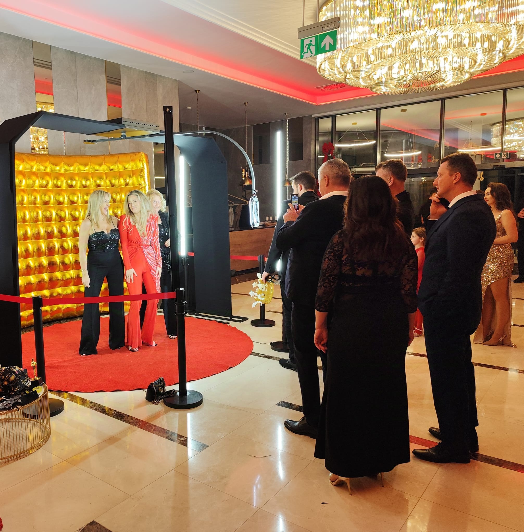 Elegancka fotobudka z złotym tłem i czerwonym dywanem. Grupa osób pozuje do zdjęcia, a kolejka gości czeka na swoją kolej. W tle widoczny kryształowy żyrandol.