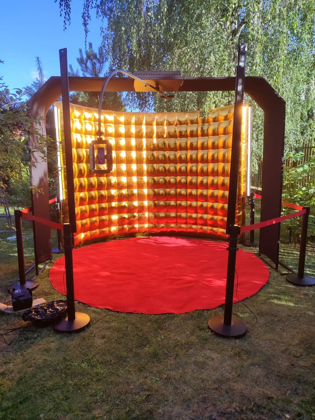 Złota, dmuchana ścianka fotobudki z czerwonym dywanem na trawie, oświetlona światłem słonecznym. Widoczne czarne słupki i barierki. Ujęcie z zewnątrz, w plenerze.