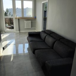 Usługi Sprzątające Joanna - Wnętrze pokoju po remoncie: szara kanapa modułowa, błyszczące płytki podłogowe, okno z widokiem na blok mieszkalny, grzejnik na ścianie.