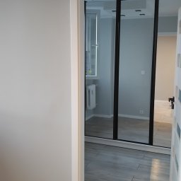 Usługi Sprzątające Joanna - Widok wnętrza mieszkania przez otwarte drzwi, ukazujący szare ściany, drewnopodobną podłogę i przesuwne drzwi do łazienki z widocznym grzejnikiem.