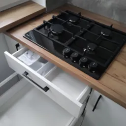 Nowoczesna kuchnia z drewnianym blatem, czarną płytą gazową Amica i otwartą białą szufladą z organizerem i instrukcją montażu.