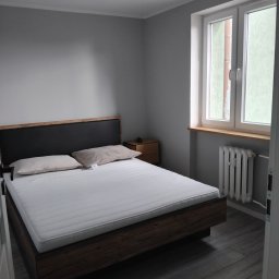 Usługi Sprzątające Joanna - Wnętrze pokoju z łóżkiem małżeńskim z drewnianą ramą i czarnym zagłówkiem, widok na szare ściany, drewnianą podłogę, białe okno z grzejnikiem pod spodem i szafkę nocną.