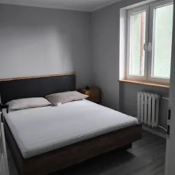 Wnętrze pokoju z łóżkiem małżeńskim z drewnianą ramą i czarnym zagłówkiem, widok na szare ściany, drewnianą podłogę, białe okno z grzejnikiem pod spodem i szafkę nocną.