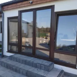 Elewacja domu z dużymi, czystymi oknami w brązowych ramach, odbijającymi otoczenie i niebo, z kamiennymi schodami prowadzącymi do wejścia.