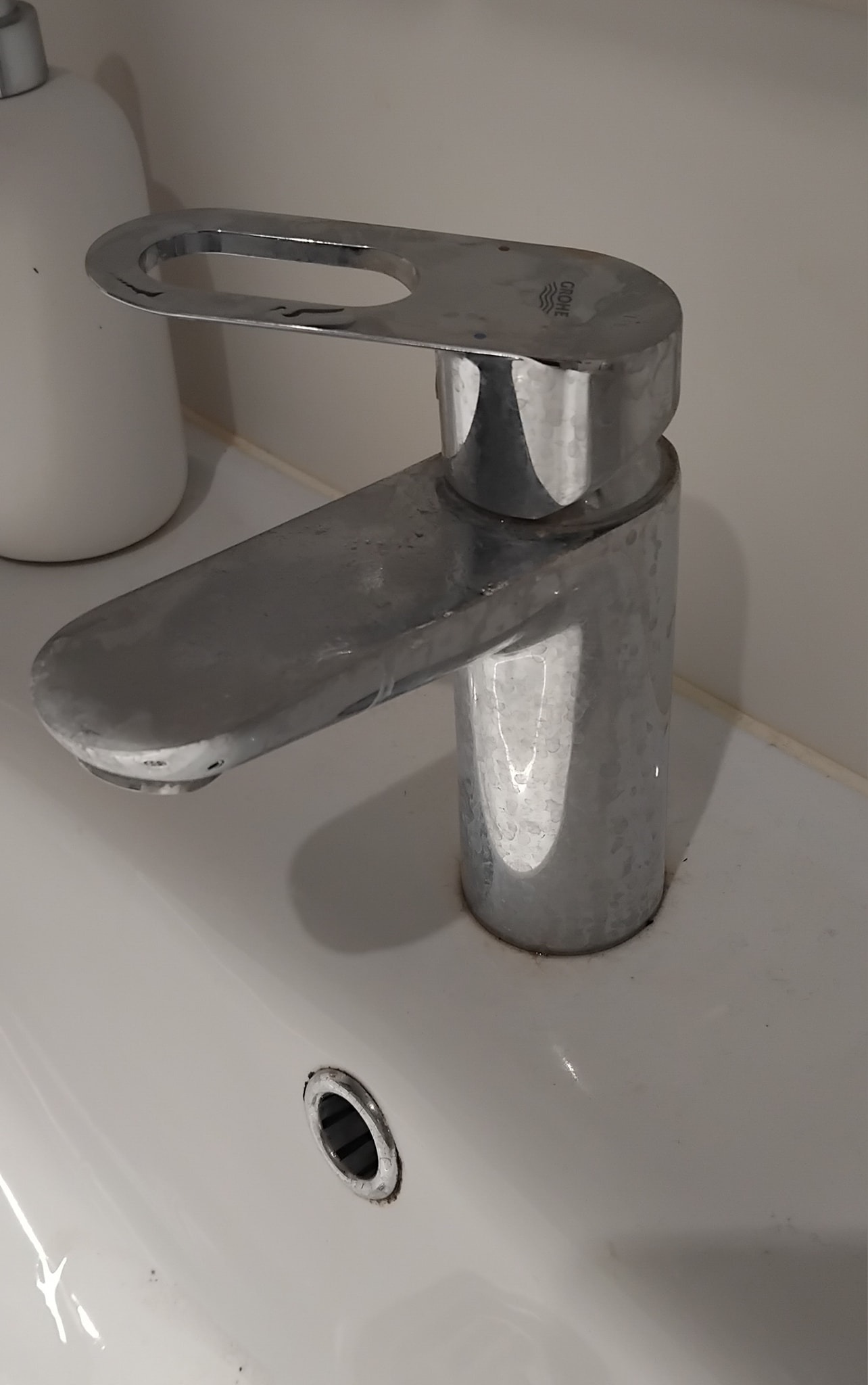 Bateria umywalkowa Grohe, pokryta zaciekami z wody i osadami, stojąca na białej umywalce z widocznym odpływem.