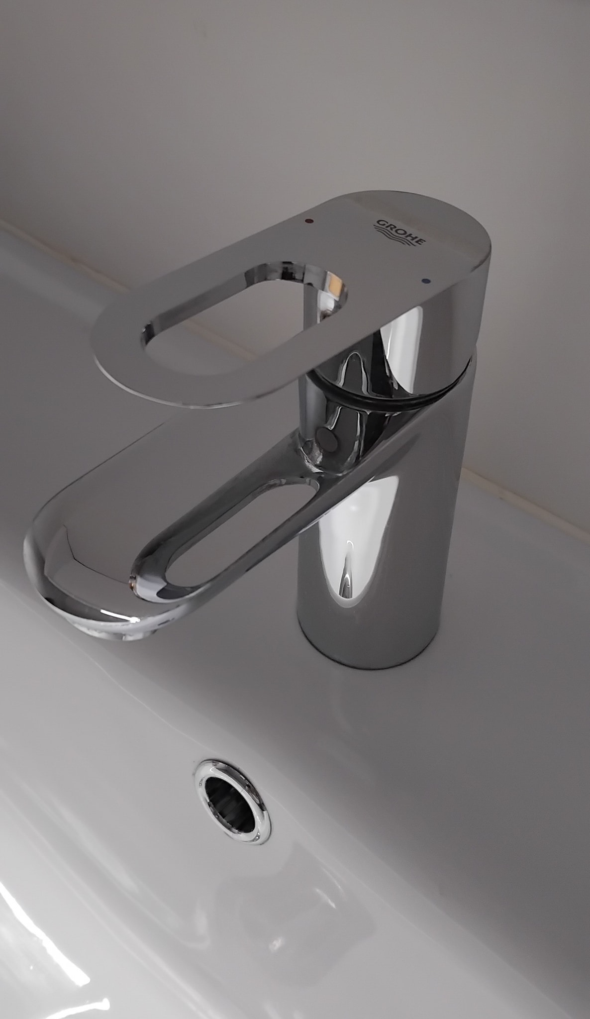 Błyszcząca, chromowana bateria umywalkowa marki Grohe zamontowana na białej umywalce, widoczny otwór odpływowy.