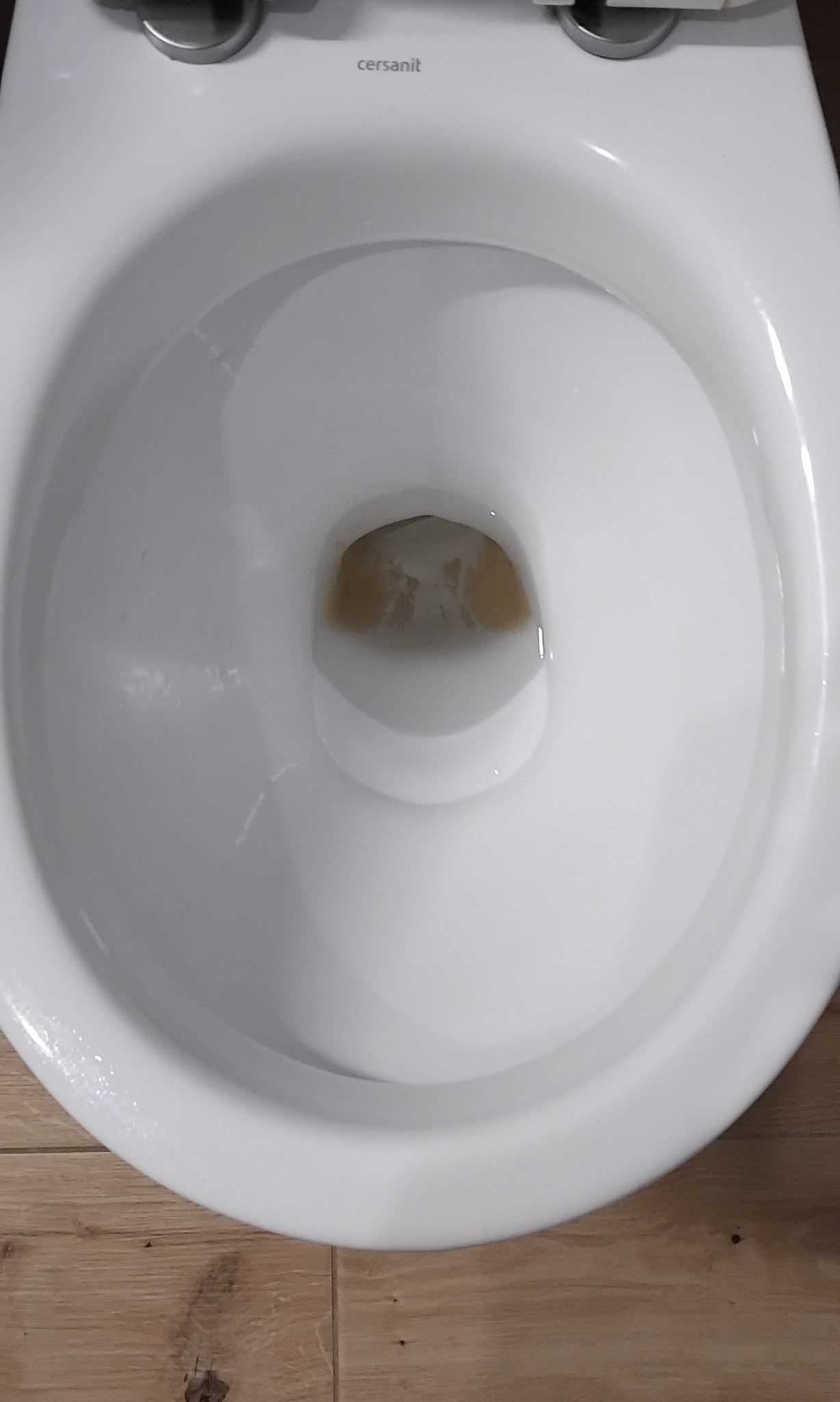 Wnętrze brudnej, białej muszli klozetowej marki Cersanit z widocznymi osadami i plamami na dnie, stojącej na drewnianej podłodze.