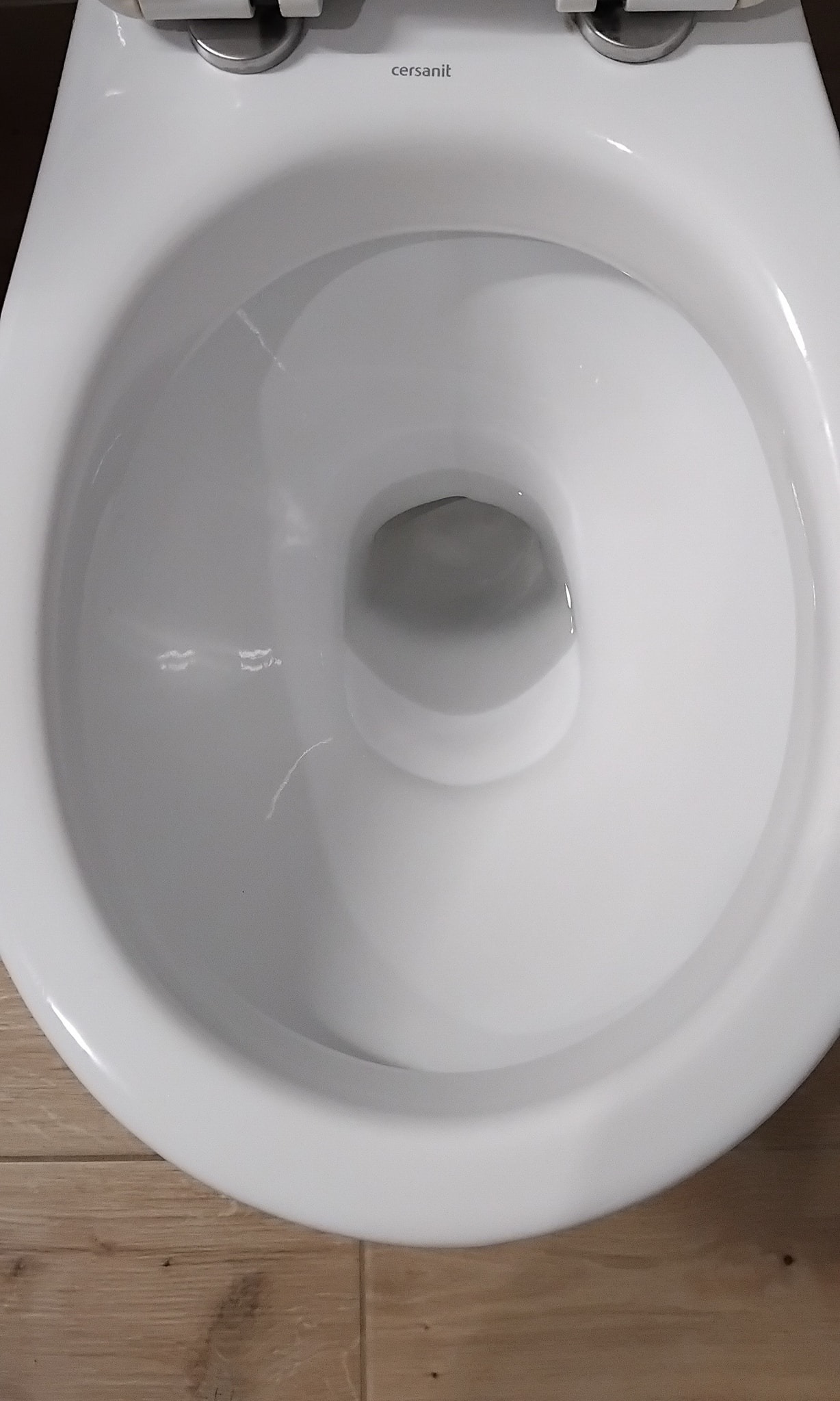 Biała, ceramiczna muszla klozetowa marki Cersanit, widok z góry, po dokładnym umyciu, z widocznymi płytkami podłogowymi imitującymi drewno.