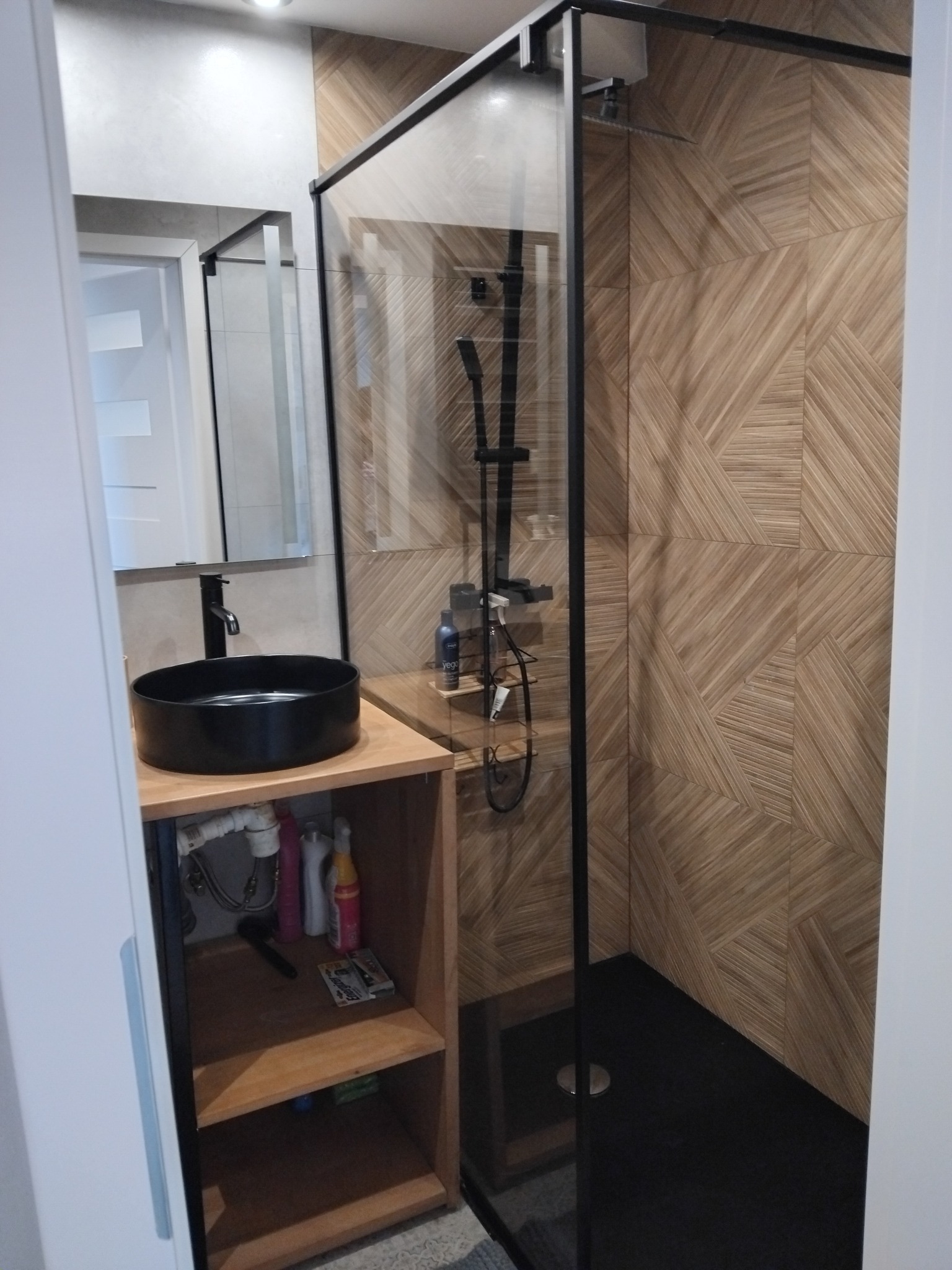 Nowoczesna łazienka z czarną umywalką nablatową, kabiną prysznicową typu walk-in z czarnymi profilami i drewnopodobnymi płytkami na ścianach.