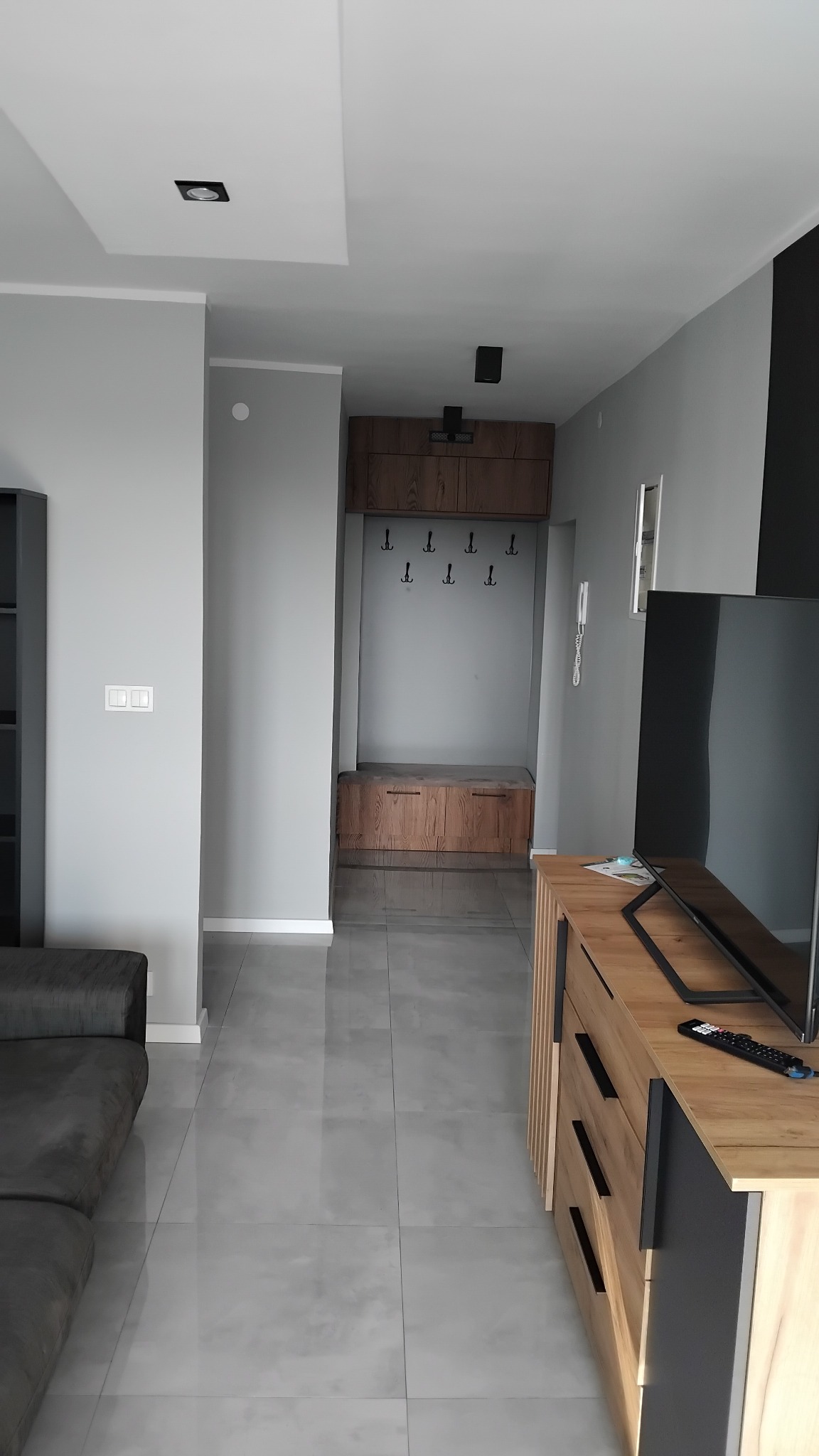 Nowoczesny przedpokój z szarymi płytkami podłogowymi, drewnianą zabudową z wieszakami i szafką na buty, widok z salonu z fragmentem szarej sofy i drewnianej komody z telewizorem.