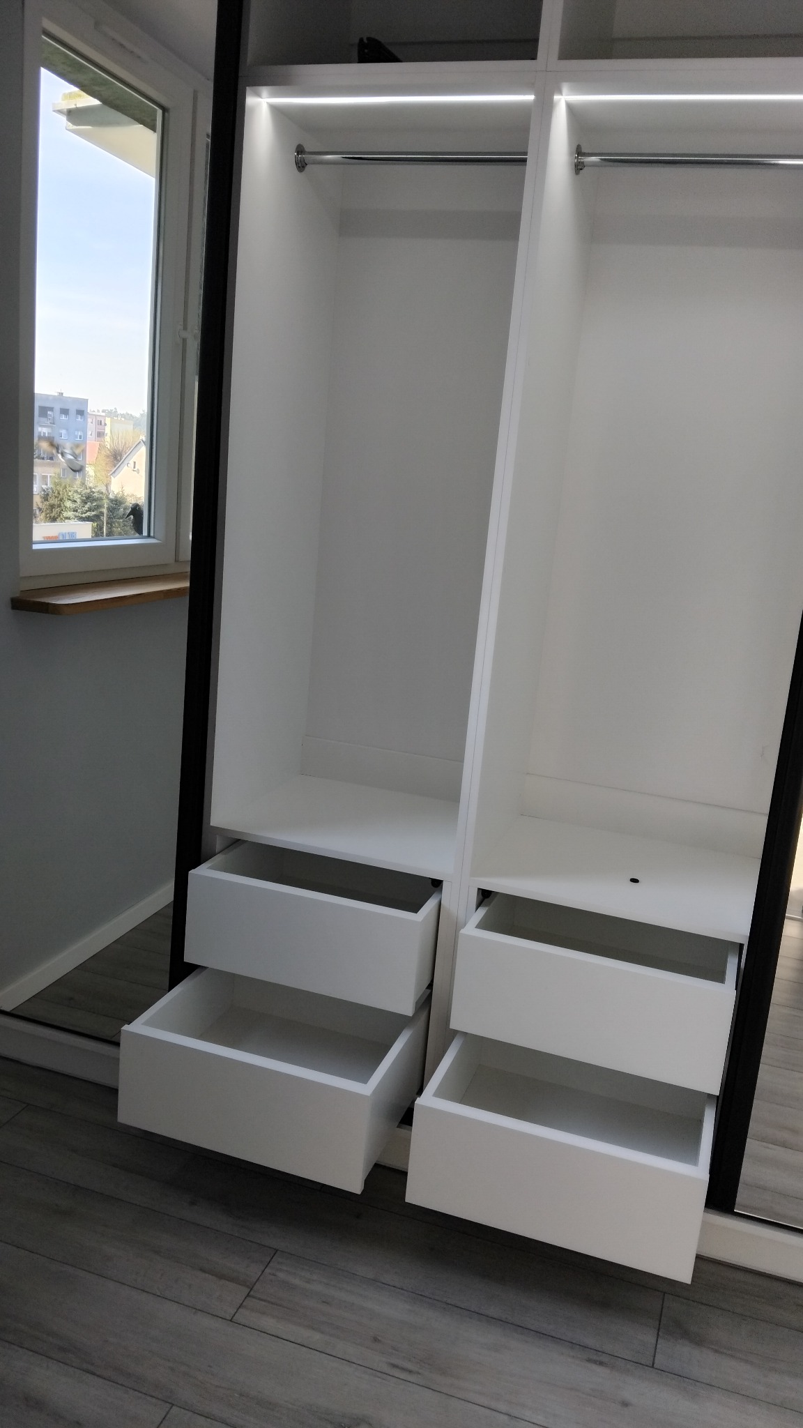 Biała, otwarta szafa wnękowa z szufladami i oświetleniem LED, widok z boku na tle okna.