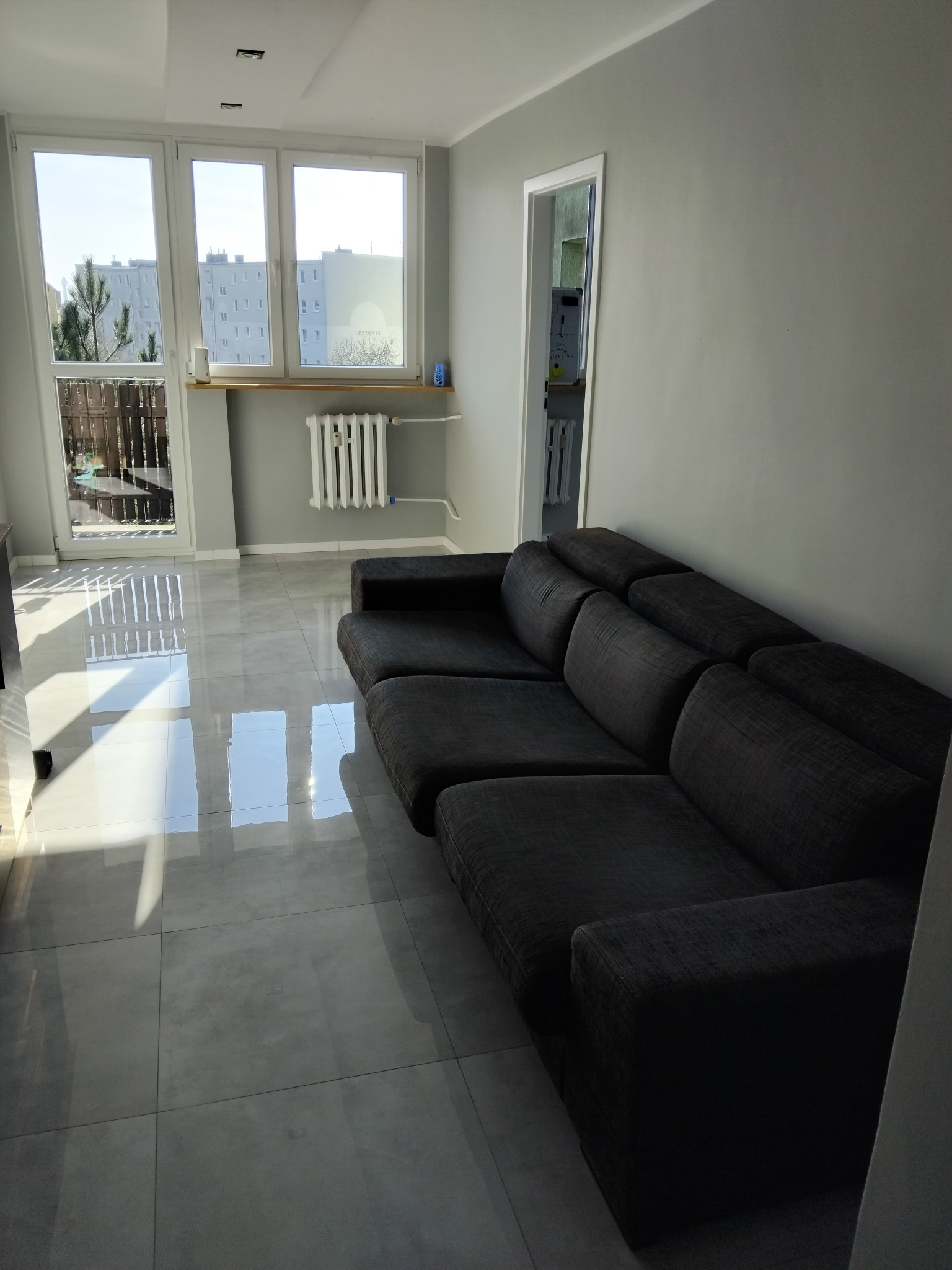 Wnętrze pokoju po remoncie: szara kanapa modułowa, błyszczące płytki podłogowe, okno z widokiem na blok mieszkalny, grzejnik na ścianie.