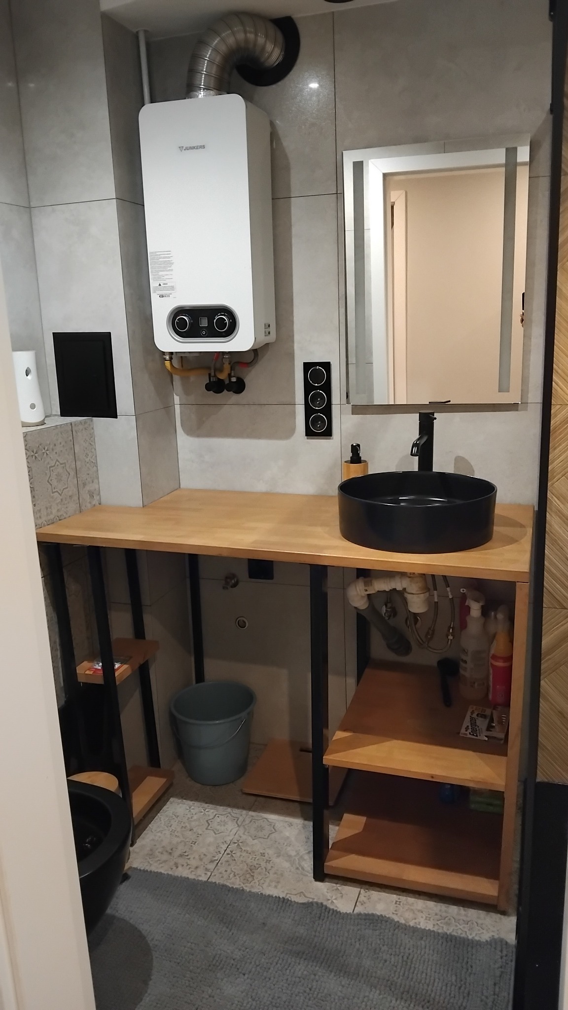 Nowoczesna łazienka z umywalką nablatową w kolorze czarnym, drewnianym blatem i szafką z otwartymi półkami, na ścianie wiszący podgrzewacz wody Junkers i lustro z oświetleniem LED.
