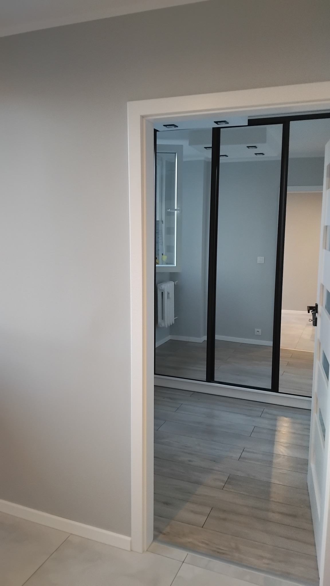 Widok wnętrza mieszkania przez otwarte drzwi, ukazujący szare ściany, drewnopodobną podłogę i przesuwne drzwi do łazienki z widocznym grzejnikiem.