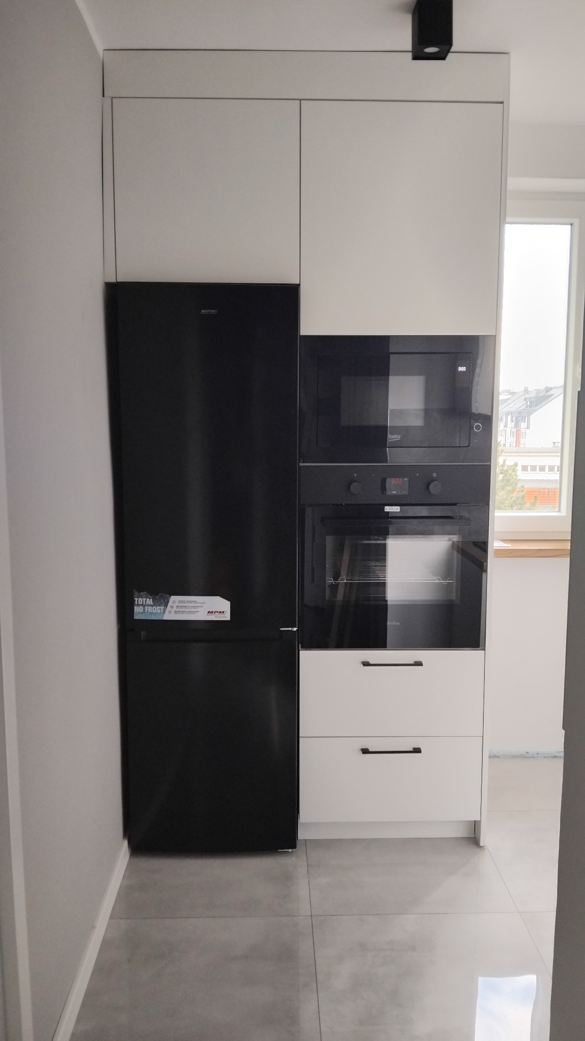 Czarna lodówka MPM obok zabudowanej szafki z piekarnikiem Amica i mikrofalówką Beko w nowoczesnej, jasnej kuchni z szarymi płytkami podłogowymi.
