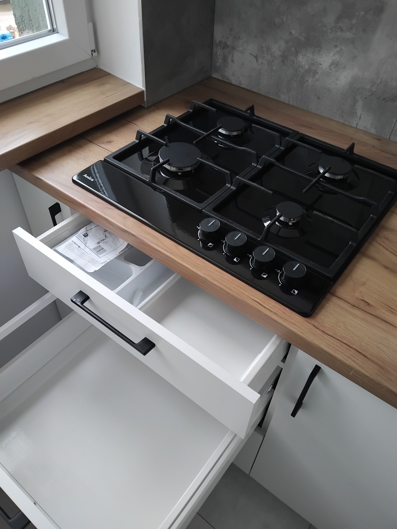 Nowoczesna kuchnia z drewnianym blatem, czarną płytą gazową Amica i otwartą białą szufladą z organizerem i instrukcją montażu.