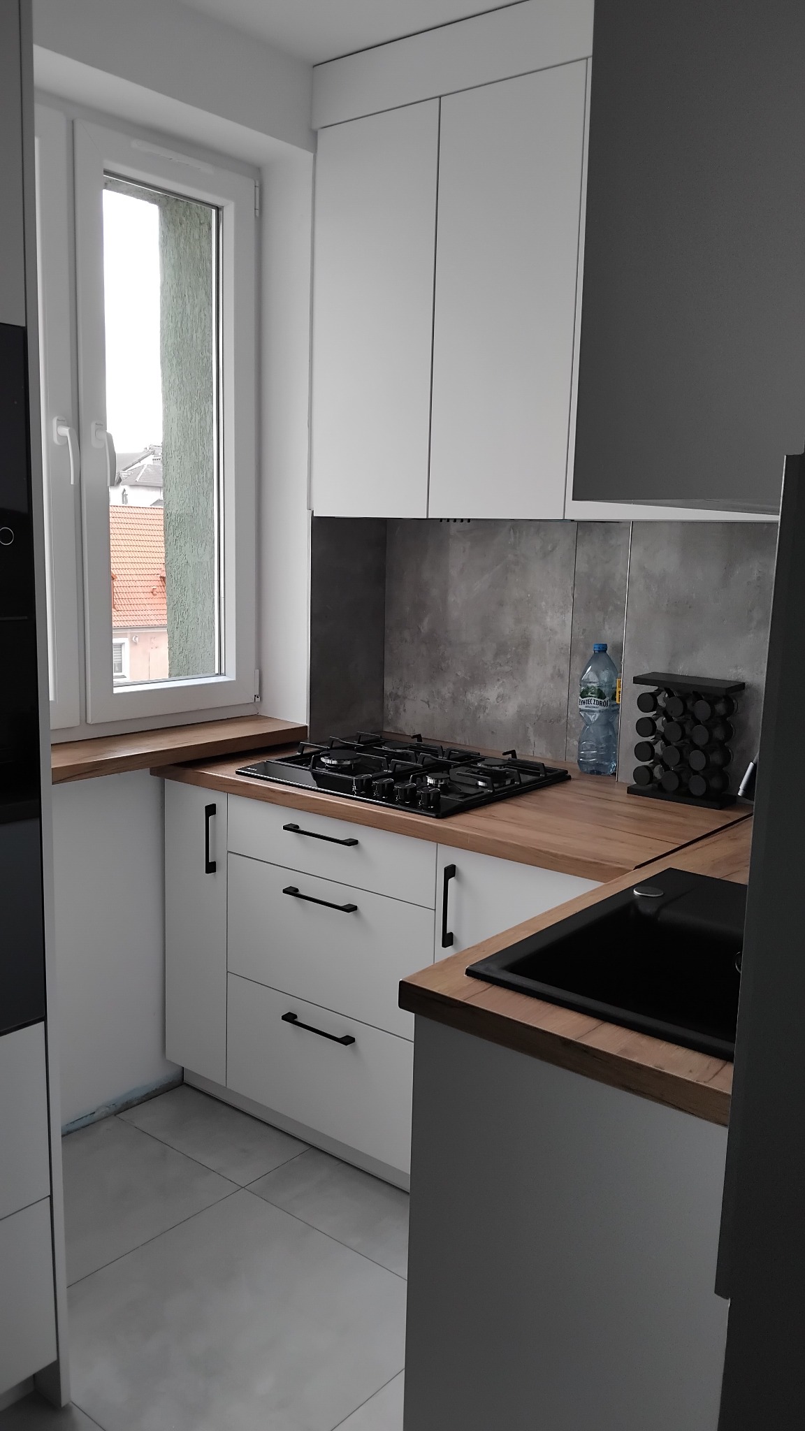 Nowoczesna kuchnia z białymi szafkami, drewnianym blatem, czarnym zlewem i płytą gazową. Widoczna butelka wody mineralnej i stojak na przyprawy. Za oknem widok na budynki mieszkalne.
