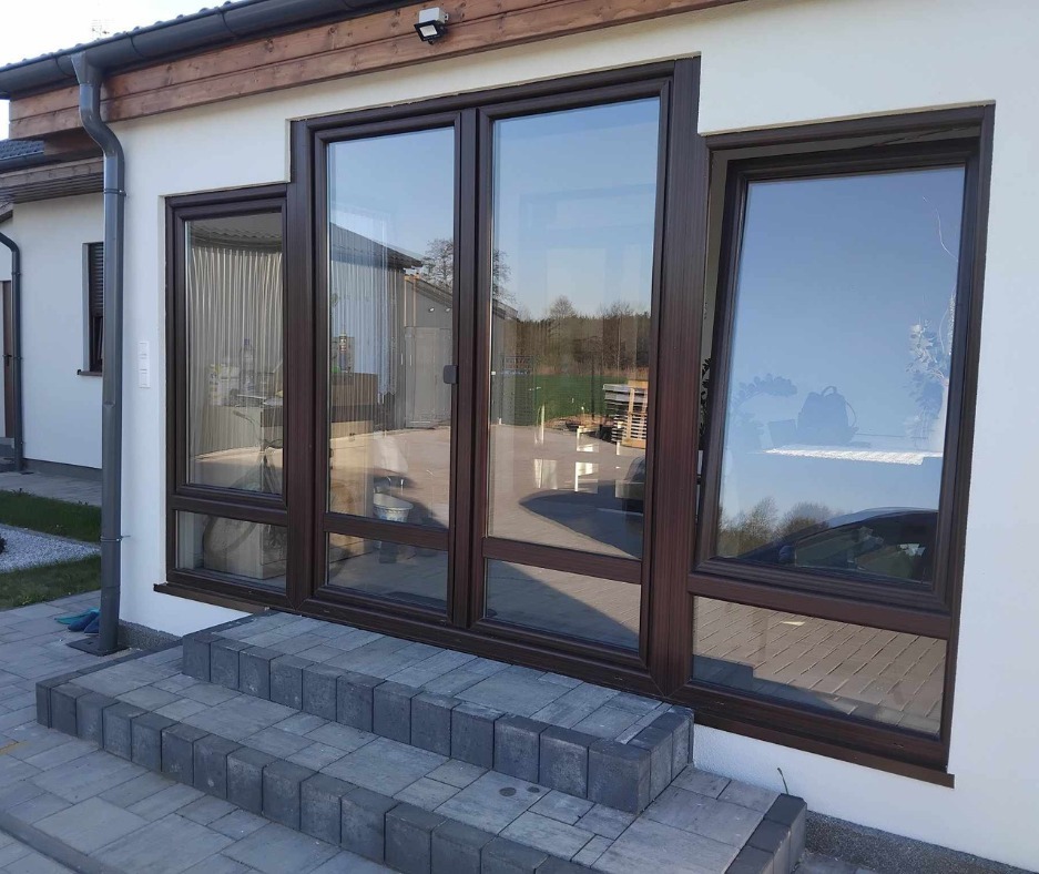 Elewacja domu z dużymi, czystymi oknami w brązowych ramach, odbijającymi otoczenie i niebo, z kamiennymi schodami prowadzącymi do wejścia.