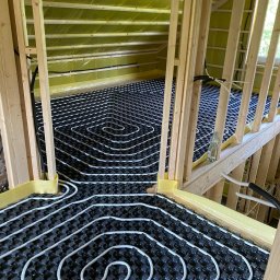 GREEN ENERGY- Nowoczesne Techniki Grzewcze Krystian Dorosz - Instalacja ogrzewania podłogowego w nowym budynku, widoczne białe rury ułożone na czarnej macie izolacyjnej, drewniana konstrukcja ścian i poddasza, żółta folia izolacyjna na ścianach, butelka wody...