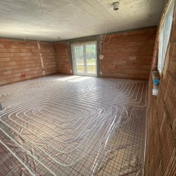 GREEN ENERGY- Nowoczesne Techniki Grzewcze Krystian Dorosz - Rozłożone rury ogrzewania podłogowego na siatce w surowym wnętrzu z ceglanymi ścianami i betonowym sufitem, przygotowane do zalania wylewką.