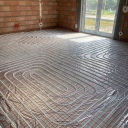 GREEN ENERGY- Nowoczesne Techniki Grzewcze Krystian Dorosz - Rury ogrzewania podłogowego ułożone na folii izolacyjnej z czerwoną siatką w surowym wnętrzu z ceglanymi ścianami i dużym oknem.