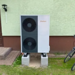 Zewnętrzna jednostka pompy ciepła Vaillant aroTHERM zamontowana na betonowych bloczkach przed budynkiem z zieloną elewacją i brązowym cokołem, obok zaparkowany rower.