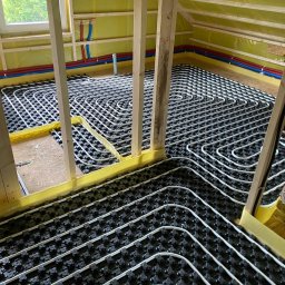 GREEN ENERGY- Nowoczesne Techniki Grzewcze Krystian Dorosz - Widok na instalację ogrzewania podłogowego w trakcie montażu, rury w systemie rozprowadzonym na czarnej macie, widoczne elementy konstrukcji drewnianej poddasza i izolacja żółtą folią.