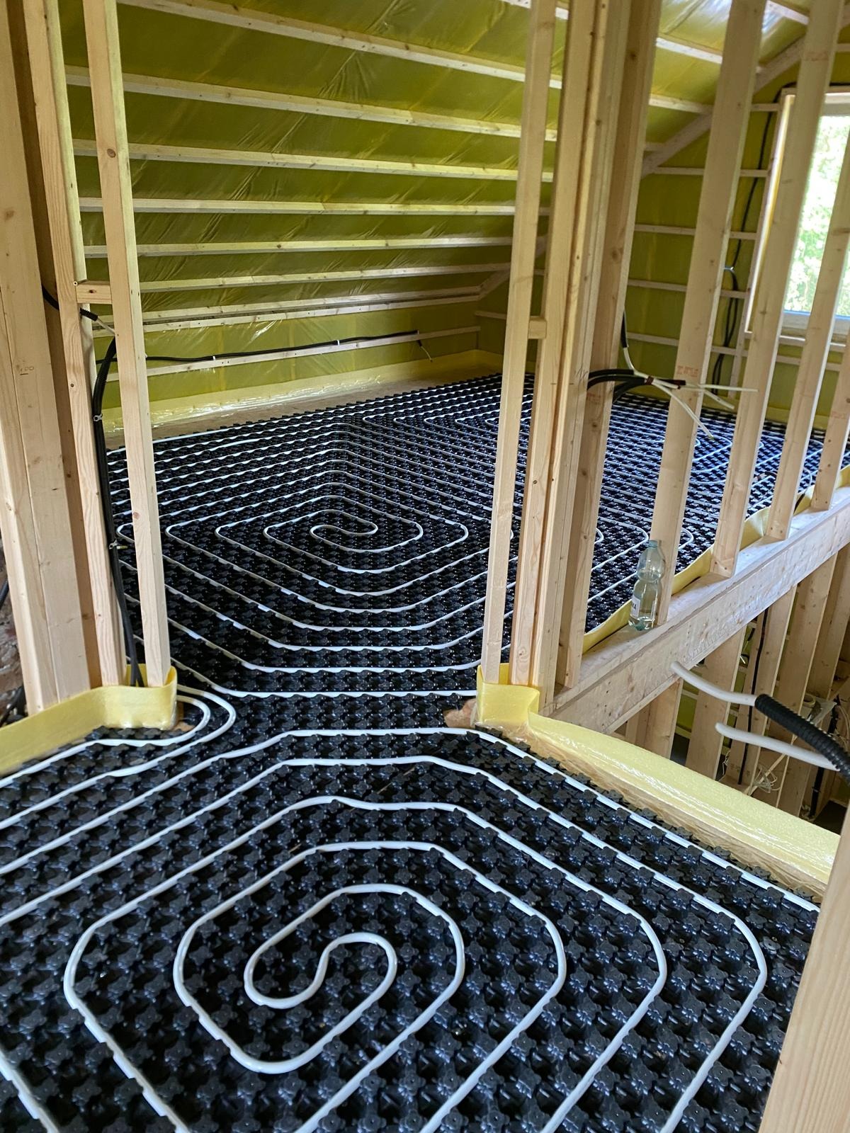 Instalacja ogrzewania podłogowego w nowym budynku, widoczne białe rury ułożone na czarnej macie izolacyjnej, drewniana konstrukcja ścian i poddasza, żółta folia izolacyjna na ścianach, butelka wody...