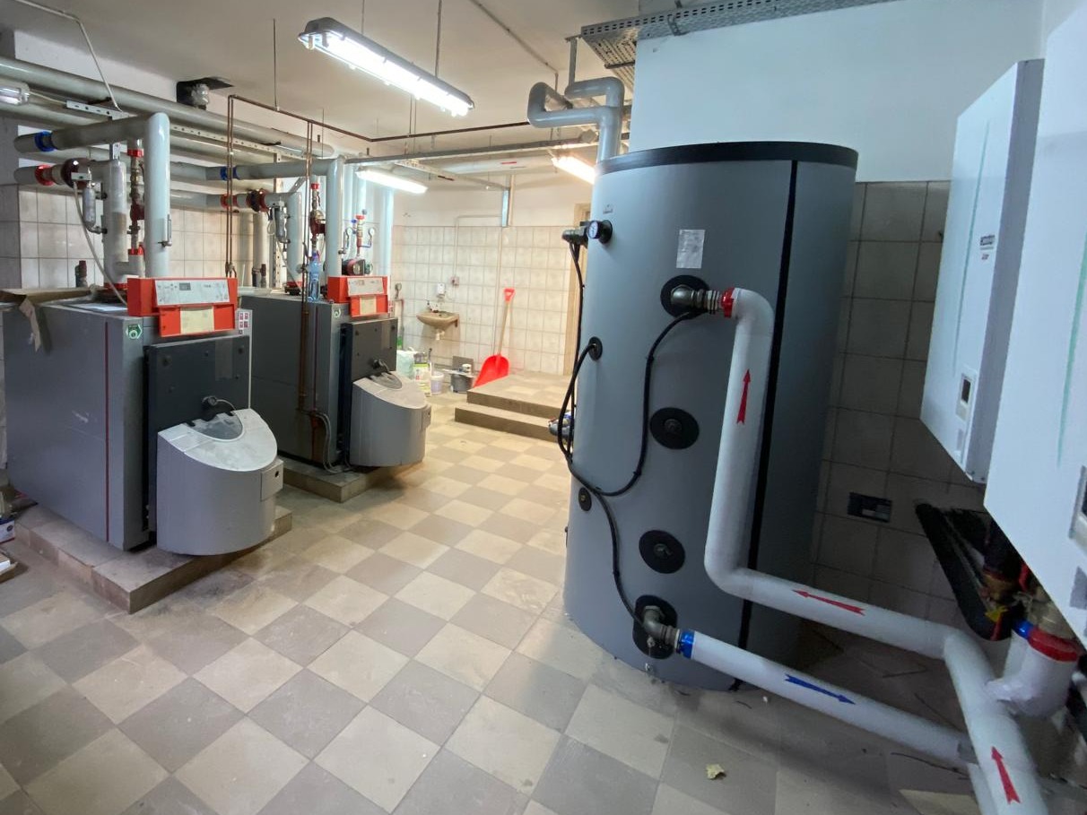 Dwa szare kotły grzewcze z pomarańczowymi akcentami i zbiornik buforowy w kotłowni z wykafelkowanymi ścianami i podłogą w szachownicę.