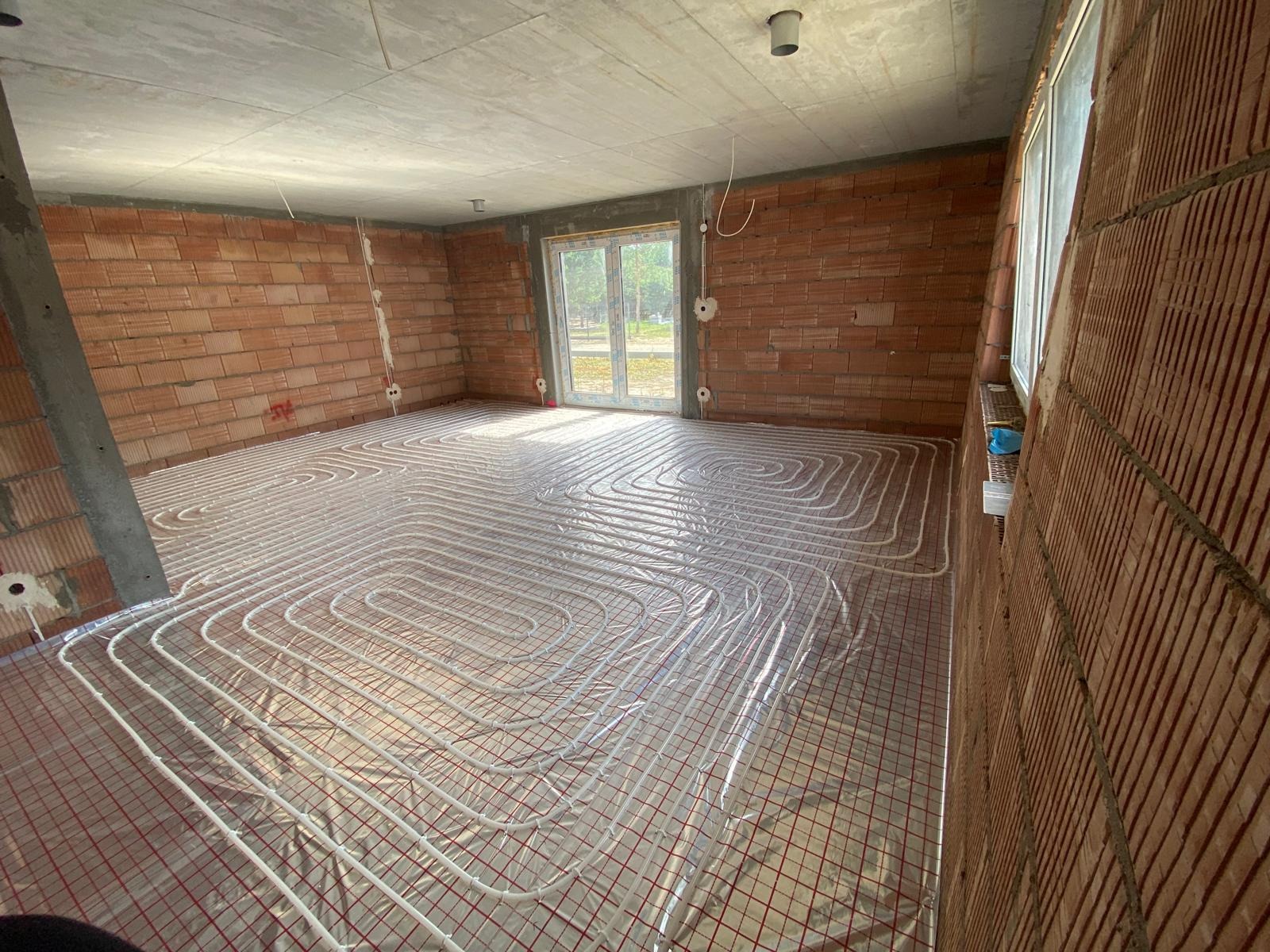 Rozłożone rury ogrzewania podłogowego na siatce w surowym wnętrzu z ceglanymi ścianami i betonowym sufitem, przygotowane do zalania wylewką.