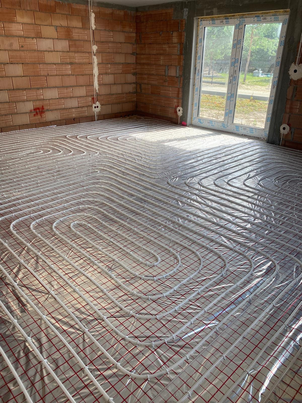 Rury ogrzewania podłogowego ułożone na folii izolacyjnej z czerwoną siatką w surowym wnętrzu z ceglanymi ścianami i dużym oknem.
