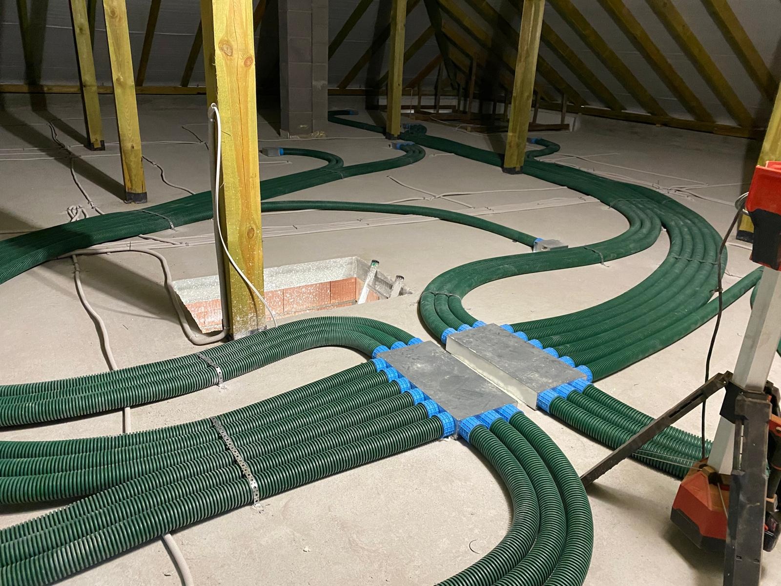 Rozprowadzenie zielonych, karbowanych rur wentylacyjnych z niebieskimi złączkami na poddaszu, widoczne drewniane elementy konstrukcji dachu i betonowe bloczki.