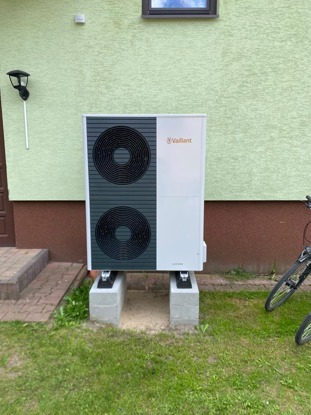 Zewnętrzna jednostka pompy ciepła Vaillant aroTHERM zamontowana na betonowych bloczkach przed budynkiem z zieloną elewacją i brązowym cokołem, obok zaparkowany rower.