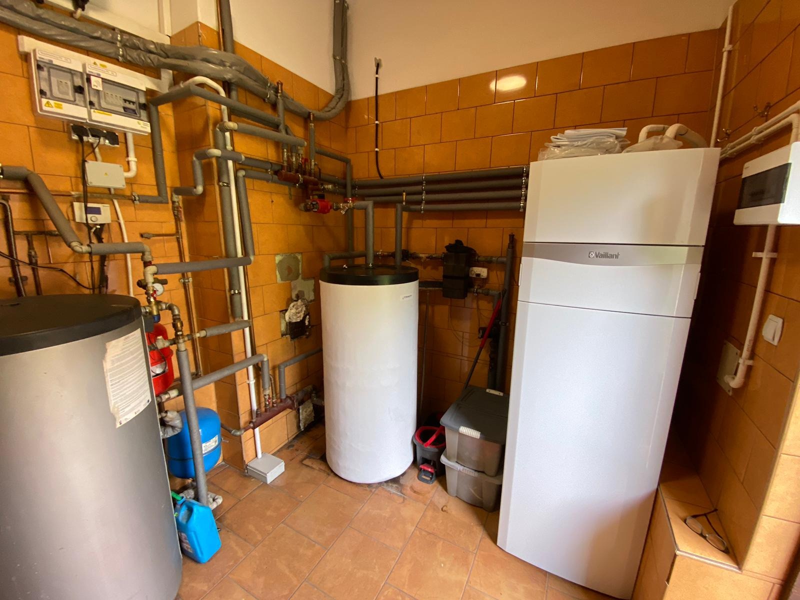 Kotłownia z instalacją centralnego ogrzewania, widoczne rury, zbiorniki, armatura i piec Vaillant na tle pomarańczowych płytek.