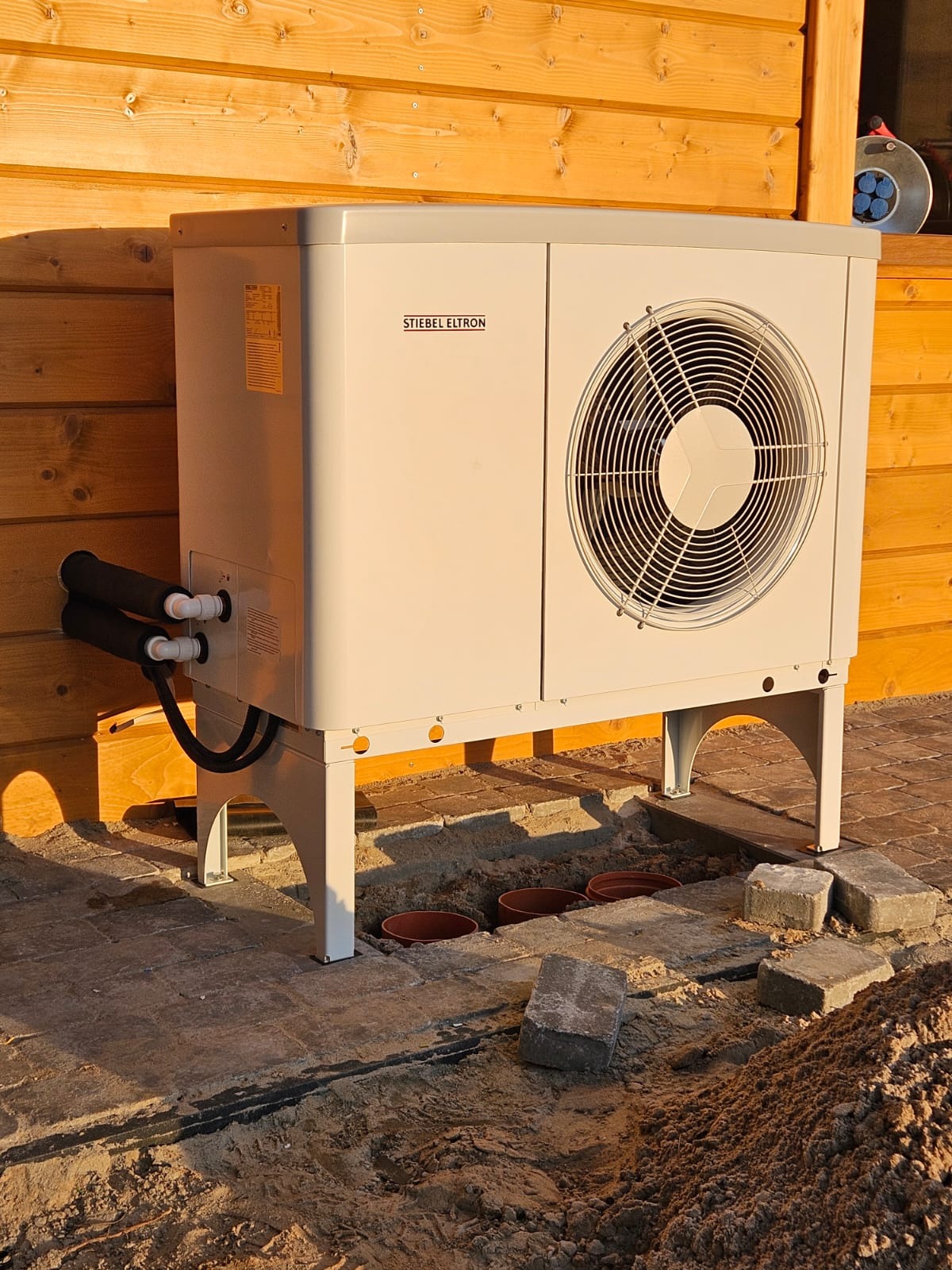 Zewnętrzna jednostka pompy ciepła Stiebel Eltron zamontowana przy drewnianej elewacji budynku, widoczne przyłącza i przygotowane podłoże z kostki brukowej z miejscami na doniczki.