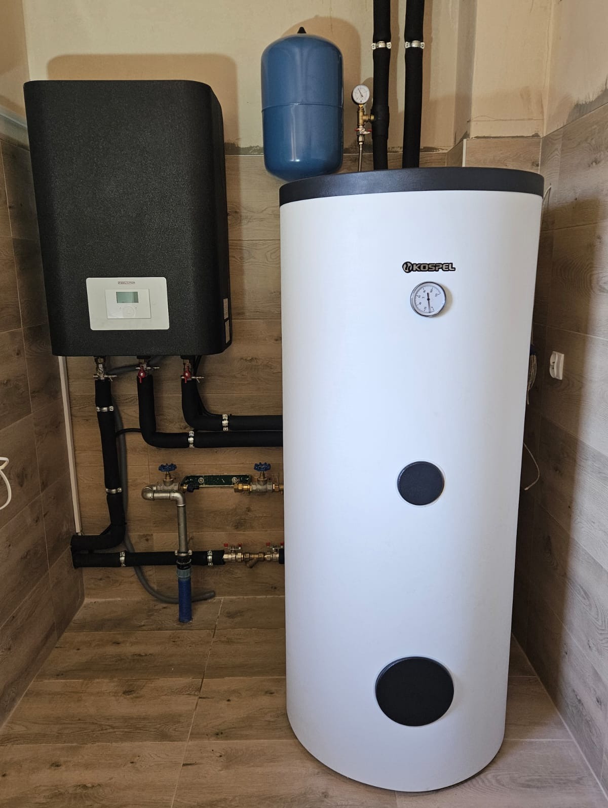 Zamontowany zestaw grzewczy Kospel z czarnym modułem sterującym i białym zasobnikiem ciepłej wody, rury w izolacji, manometr na ścianie, płytki imitujące drewno na podłodze i ścianach.